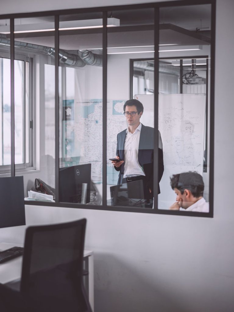 Deux personnes dans un bureau, l’une assise et l’autre debout devant des ordinateurs portables, vues à travers une fenêtre vitrée